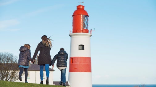 Three people walk away from the camera towards a red and white striped lighthouse